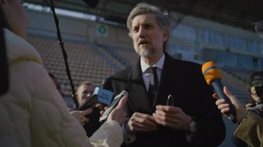 Organizasyon temsilcisi basın sorularını yanıtlıyor, futbol stadyumunun dışında röportaj veriyor. TV haberleri için basın toplantısında özel yatırımcıyla röportaj. Basın kampanyası sırasında gazeteciler kalabalık.