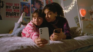 Yatak odasında sohbet eden iki çok kültürlü arkadaş ve cep telefonu kullanarak komik selfie çekimleri. Mutlu genç kızlar boş zamanlarını yatakta yatarak geçirirler. Arkadaşlar birlikte eğleniyorlar..