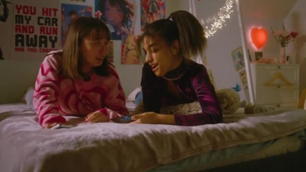 卧室里的两个多文化少女一边笑着一边说话 一边用手机看有趣的视频 快乐的少女在家里躺在床上消磨时光 最好的朋友在一起玩 — 图库视频影像