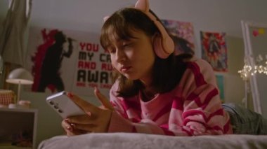 Genç kız, rahat yatak odasındaki yatakta uzanan cep telefonuyla kulaklıkla müzik dinliyor ve sosyal medyayı kaydırıyor. Asyalı kız evde eğlenip eğleniyor. Yaşam tarzı kavramı.