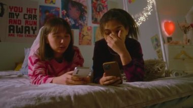 Yatak odasında sohbet eden iki çok kültürlü arkadaş ve cep telefonu kullanarak komik selfie çekimleri. Mutlu genç kızlar boş zamanlarını yatakta yatarak geçirirler. Arkadaşlar birlikte eğleniyorlar..