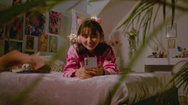 Kulaklıklı Asyalı genç kız görüntülü sohbet yoluyla konuşuyor, rahat yatak odasında yatakta yatarken cep telefonu kullanarak internette geziniyor. Kızlar boş zamanlarını evde geçirip eğleniyorlar. Yaşam tarzı kavramı.