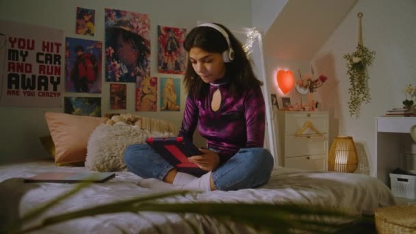 戴着耳机的美丽的非洲裔美国女孩坐在自己房间的床上 用平板电脑上网 青少年快乐地和朋友聊天或听音乐 舒适舒适的家 内部舒适时尚 — 图库视频影像
