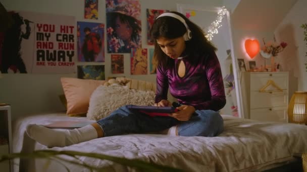 ヘッドフォンの美しいティーンエイジャーは 彼女の部屋のベッドに座り タブレットを使用してインターネットをサーフ アフリカ系アメリカ人の女の子はビデオコンテンツを見たり 映画を見たりします 居心地の良いスタイリッシュなインテリアで快適な家 — ストック動画