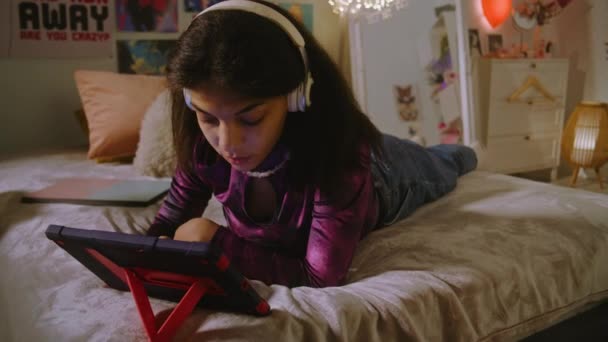 戴耳机的漂亮少女躺在房间的床上 用平板电脑上网 非裔美国女孩与朋友聊天或观看视频内容 在舒适的家中度过闲暇时间 — 图库视频影像