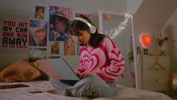 戴耳机的少女坐在舒适的卧室的床上 一边听音乐 一边聊天 一边在笔记本电脑上滚动社交网络 蒙古女孩享受周末 在家里玩得很开心 生活方式概念 — 图库视频影像