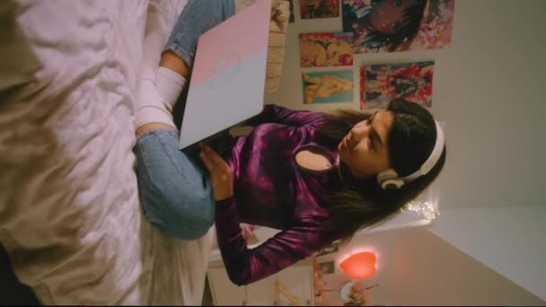 一个少女在耳机里与朋友在线聊天 在笔记本电脑上上网的垂直视频坐在卧室的床上 非洲裔美国女孩在家里玩得很开心 — 图库视频影像