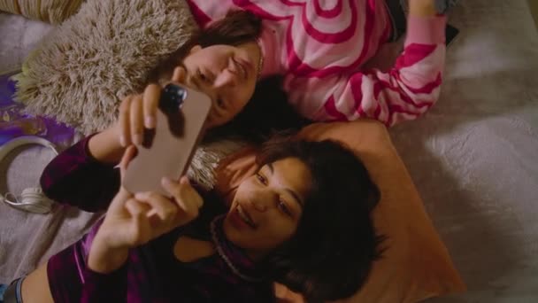 两个不同的好友在舒适的卧室里笑着 用智能手机自嘲一番 快乐的少女在家里躺在床上消磨时光 快乐的少女在一起玩乐 — 图库视频影像