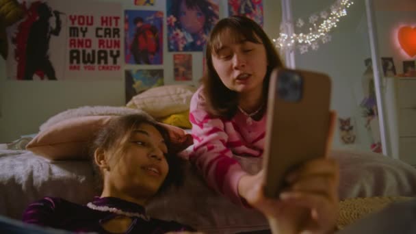 アフリカ系アメリカ人の女の子が床に座り 電話を使ってインターネットをサーフ モンゴル人のティーンはベッドに横たわり 友人とのコンテンツを見ています 複数の民族の女の子が一緒に家でレジャー時間を過ごしています 友人関係 — ストック動画