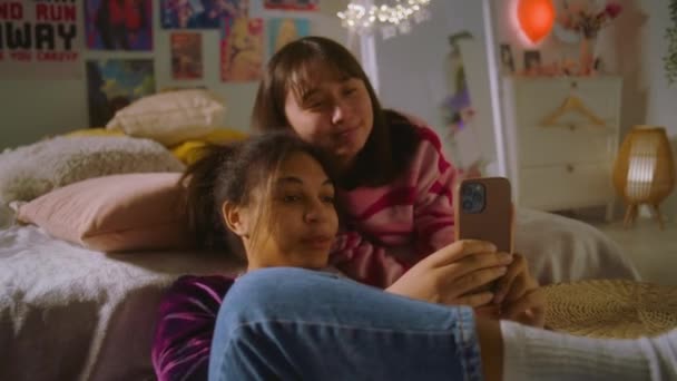 非洲裔美国女孩坐在地板上 用电话上网 亚洲少年躺在床上 在电话旁观看 两个多民族女孩在一起用电话拍照 朋友关系 — 图库视频影像