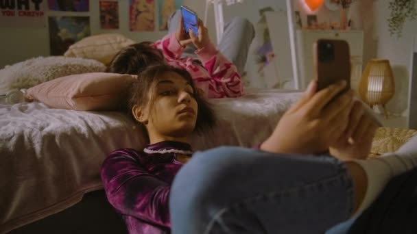 複数の民族の女の子が一緒に家でレジャー時間を過ごしています アフリカ系アメリカ人の女の子がベッドの近くに座り 電話でインターネットを使っている モンゴル人のティーンは友人に会い 彼女の携帯を見る 友人関係 — ストック動画