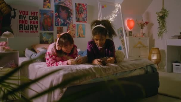 寝室で2人の多民族の十代の少女が スマートフォンでインターネットを閲覧したり ヘッドフォンで音楽を聴いたりしながら笑ったりします ベストガールフレンドは ベッドの上に横たわって自宅で一緒に楽しんでいます — ストック動画