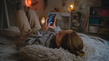 Kulaklıklı genç bir kız evdeki yatakta uzanıyor, müzik dinliyor ve uygulamadan cep telefonu kullanarak şarkı seçiyor. Beyaz tenli genç, boş zamanlarını gündüzleri rahat yatak odasında geçirir. Yaşam tarzı kavramı.