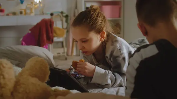 Young girl lies on bed, eats croissant, watches video content using tablet. Boy lies near sister and uses phone. Teenagers spend leisure time together. Family relationship. Home with stylish interior.