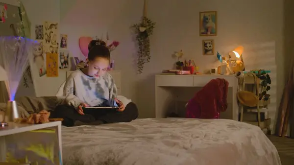Young girl sits on bed in her room and surfs the internet or chats online using tablet. Cute schoolgirl spending leisure time at home scrolling social networks. Home with cozy and stylish interior.