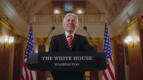 美国总统站在讲台的后面 发表了富有启发性的政治演讲 年长的政治家或部长在白宫的新闻发布会上发表讲话 背景中的美国国旗 — 图库视频影像