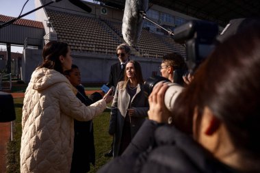 İki organizasyon temsilcisi basın sorularını yanıtlıyor ve dışarıda bir futbol stadyumunda röportaj veriyor. TV haberleri için basın toplantısında başarılı bir SEO röportajı. Gazeteciler basın kampanyasında kalabalıklar.