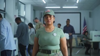 Kamuflaj üniformalı bir kadın oy kullanma merkezinde dikiliyor ve kameraya bakıyor. Kadın asker portresi, Amerika Birleşik Devletleri seçmeni. Oy verme kabinleri olan bir arka plan. Yurttaşlık görevi kavramı.