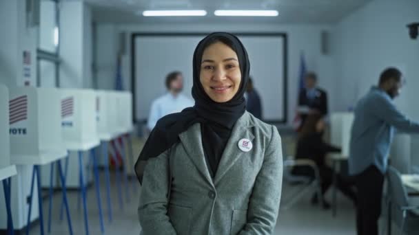 女性は現代の投票所に立って ポーズ そしてカメラを見ています ムスリム女性の肖像画 アメリカ合衆国選挙の有権者 投票ブースの背景 市民の義務に関する概念 — ストック動画