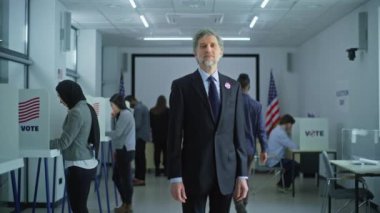 Yetişkin bir adamın portresi, Amerika Birleşik Devletleri seçmeni. İş adamı modern bir seçim merkezinde duruyor, poz veriyor ve kameraya bakıyor, gülümsüyor. Oy kullanma kabinleri ve Amerikan bayrakları olan arka plan.