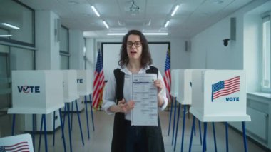 Amerikalı kadın seçmen ya da sandık memuru kamerada konuşur, kağıt oylarını gösterir, oy kullanma çağrısı yapar. Birleşik Devletler 'de Ulusal Seçim Günü. Sandık merkezinde oy kullanma kabinleri. Vatandaşlık görevi ve vatanseverlik