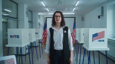 Rozetli bir Amerikan vatandaşı kameraya konuşuyor, oy verme çağrısı yapıyor. Birleşik Devletler 'de Ulusal Seçim Günü. Oy kullanma merkezinin arka planında oy kullanma kabinleri var. Vatandaşlık görevi. Dolly vurdu..
