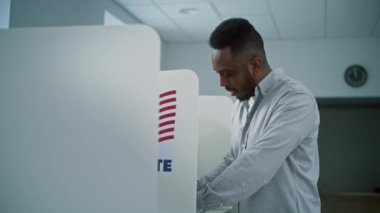 Afro-Amerikalı bir adam oy kabininde oy pusulası dolduruyor, sonra gülümsüyor ve kameraya bakıyor. Amerika Birleşik Devletleri Ulusal Seçim Günü 'nde sandık merkezinde ABD vatandaşı veya erkek seçmen.