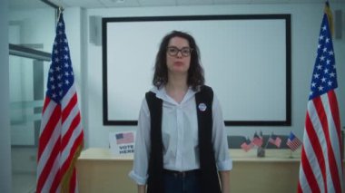 Rozeti olan Amerikalı kadın seçmen ya da sandık görevlisi kameraya konuşuyor ve oy kullanma çağrısı yapıyor. Birleşik Devletler 'de Ulusal Seçim Günü. Sandık merkezinde oy kullanma kabinleri. Vatandaşlık görevi ve vatanseverlik