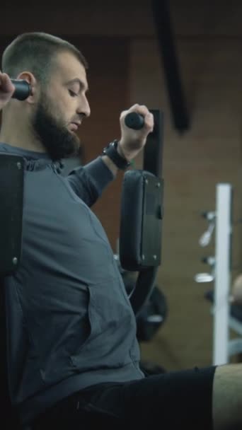 プロのスポーツジム機器を備えた現代のフィットネスセンターでトレーニングマシンに義足の列車を持つ男性の垂直ショット 別の成人アスリートは 背景で腕の演習を行っています — ストック動画