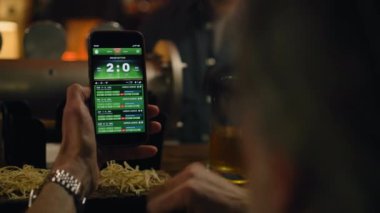 Spor taraftarlarının maç skorlarını ve cep telefonu kullanarak bahisçi reytinglerini kapat. Beyaz adam, geceleri barın barında oturarak futbol dünya ligi maçına bahis oynuyor. Kumar konsepti.