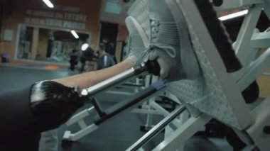 Modern spor salonunda bacak pres makinesinde protez bacak eğitimi alan motive olmuş bir sporcunun fotoğrafı. Yapay bacaklı yetişkin sporcu profesyonel spor malzemelerini kullanarak egzersiz yapıyor..