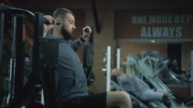 Motivasyonlu atletik adam protez bacaklı, spor aletleriyle modern fitness merkezinde egzersiz makinesine bağlanmış. Arka planda başka bir yetişkin sporcu kol egzersizi yapıyor..