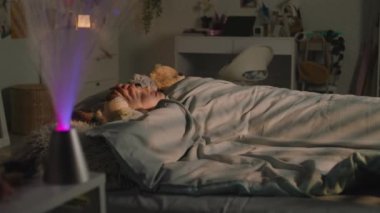 Güzel genç kız battaniyenin altında pelüş oyuncaklarla yatar ve akşamları odasında dinlenir. Sevimli liseli kız uyanıyor ve gece etrafa bakıyor. Rahat bir ev, rahat bir iç mekan..