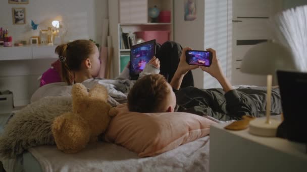 2人のティーンエイジャーが部屋のベッドに横たわり デジタルデバイスでビデオゲームをプレイする 女の子はタブレットを使用し 男の子は一緒に遊ぶために電話を使用します 姉は弟とレジャー時間を過ごした 家族関係 — ストック動画