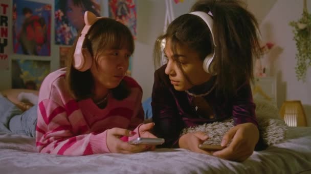 寝室で2人の多文化なティーンエイジャーがヘッドフォンで音楽を聴き 携帯電話を使ってソーシャルネットワークをスクロールしている ベッドの上に横たわる自宅で一緒に楽しんでいる親友 — ストック動画