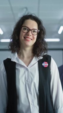 Beyaz kadın portresi, Amerika Birleşik Devletleri seçmeni. Gözlüklü kadın oy verme merkezinde duruyor, poz veriyor, gülümsüyor, kameraya bakıyor. Oy verme kabinleri olan bir arka plan. Yurttaşlık görevi kavramı.