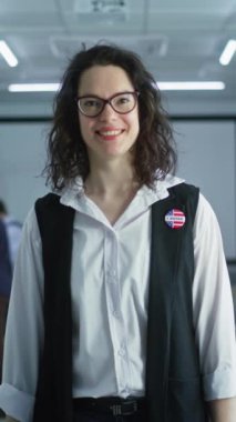 Rozetli bir kadın oy kullanma merkezinde duruyor, poz veriyor, gülümsüyor, kameraya bakıyor. Beyaz kadın portresi, Amerika Birleşik Devletleri seçmeni. Oy verme kabinleri olan bir arka plan. Yurttaşlık görevi kavramı.