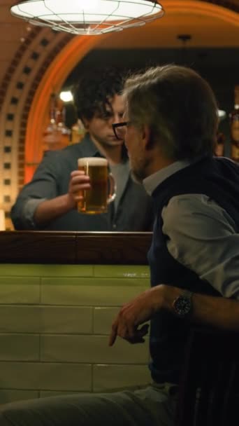 2人の男性の友人が話し 夜にスタイリッシュなパブでバーカウンターに座ってビールを飲む バーテンダーは成熟した男にビールを飲ませる 古い友人との出会い コンセプト — ストック動画