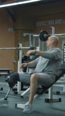 Protez bacaklı bir adamın dikey çekimi spor salonunda halterle ağırlık çalışması yapar, fitness terapisi antrenörüyle, sonra da ara verir. Yapay bacaklı sporcu sporu kullanarak egzersiz yapar.