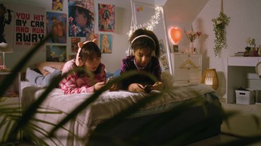 Yatak odasında çok ırklı iki genç kız, internette akıllı telefonlarıyla gezinip kulaklıkla müzik dinlerken konuşup gülüyorlar. En iyi arkadaşlar evde birlikte eğleniyor. Yatakta uzanıyorlar..