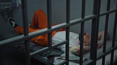 Turuncu üniformalı mahkum hücre yatağında yatıyor. Mahkum, suçtan hapis cezasına çarptırıldı. Islahevinde bir suçlu var. Adalet sistemi. Metal parmaklıklardan bak. Dolly vurdu..