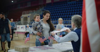 Kadın Amerikan vatandaşı oy kullanmaya sandığa geliyor. Elinde bebek olan kadın anket memuruyla konuşuyor ve bülten dinliyor. ABD başkan adaylarının siyasi ırkları. Ulusal Seçim Günü.