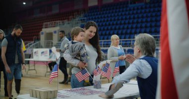 Kadın Amerikan vatandaşı oy kullanmaya sandığa geliyor. Elinde bebek olan kadın anket memuruyla konuşuyor ve bülten dinliyor. ABD başkan adaylarının siyasi ırkları. Ulusal Seçim Günü.