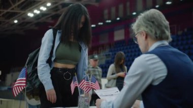 Afro-Amerikalı kız anket memuruyla konuşuyor ve bülten alıyor. Çeşitli Amerikan vatandaşları oy kullanma merkezine oy vermeye geldi. ABD başkan adaylarının siyasi ırkları. Ulusal Seçim Günü.