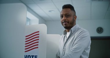 Afro-Amerikalı bir adam oy kabininde oy pusulası dolduruyor, sonra gülümsüyor ve kameraya bakıyor. Amerika Birleşik Devletleri Ulusal Seçim Günü 'nde sandık merkezinde ABD vatandaşı veya erkek seçmen.
