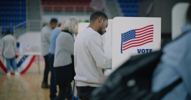 Elinde bültenle yaşlı erkek seçmen oy verme kabinine geliyor. Çok kültürlü Amerikan vatandaşları oy vermeye sandığa geliyor. ABD başkan adaylarının siyasi ırkları. Ulusal Seçim Günü.