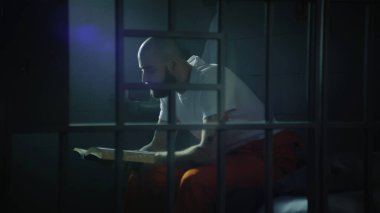 Turuncu üniformalı bir erkek mahkum hapishane hücresinde yatağında oturup İncil okuyor. Yasadışı hüküm giymiş bir adam hapiste hapis yatıyor. Metal parmaklıklardan bak. Gözaltı merkezi ya da ıslah evi.