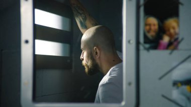 Reflection of male prisoner in mirror looking out the window in prison cell. Photos of family hang near the mirror. Inmate serves imprisonment term in jail. Detention center or correctional facility. clipart