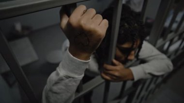 Depresyondaki Afro-Amerikan genç suçlu hapisteki hücrede duruyor, metal parmaklıklar tutuyor. Genç mahkumlar arka planda yatakta otururlar. Çocuk ıslahevi ya da ıslah evi. Yüksek açı.