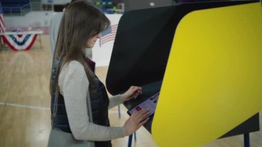 ZAPORIZHYA, UKRAINE - 15 Mart 2024: Kafkas kadın, kadın seçmen düşünüyor ve seçim sandığında oy verme kabininin yanında duruyor. ABD vatandaşı oy vermek için tablet bilgisayar kullanıyor. Başkanlık